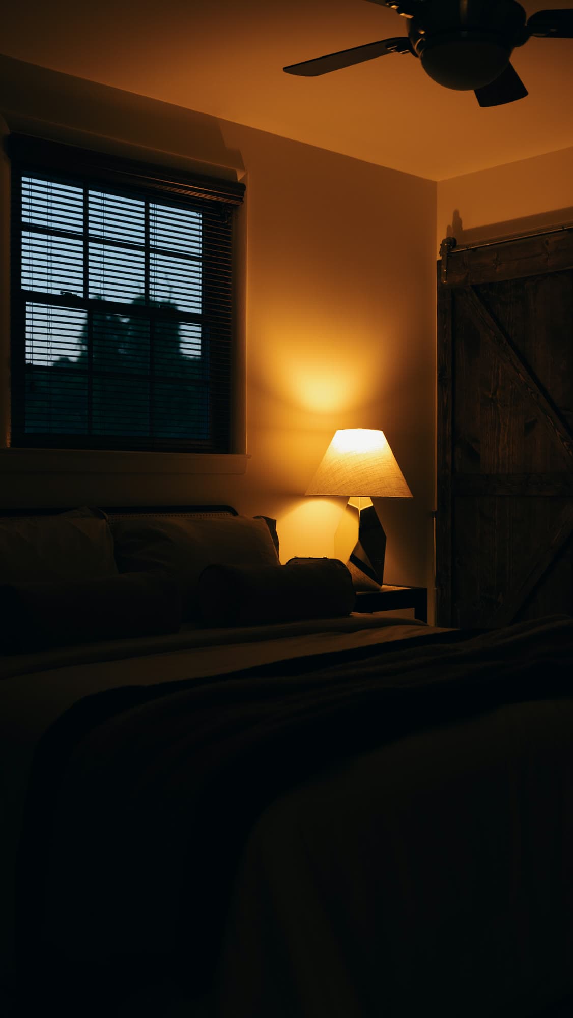 A bedside lamp washing a wall in amber light beside a window at dusk