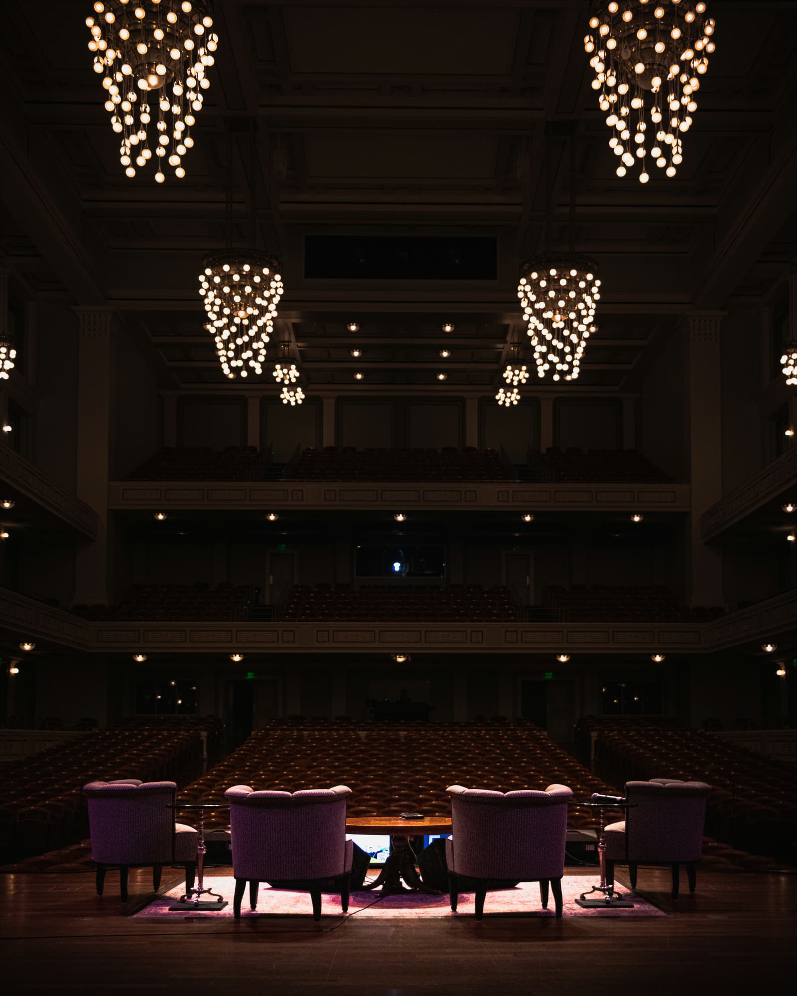 Four armchairs set for a talk on an empty concert hall stage, under glowing chandeliers