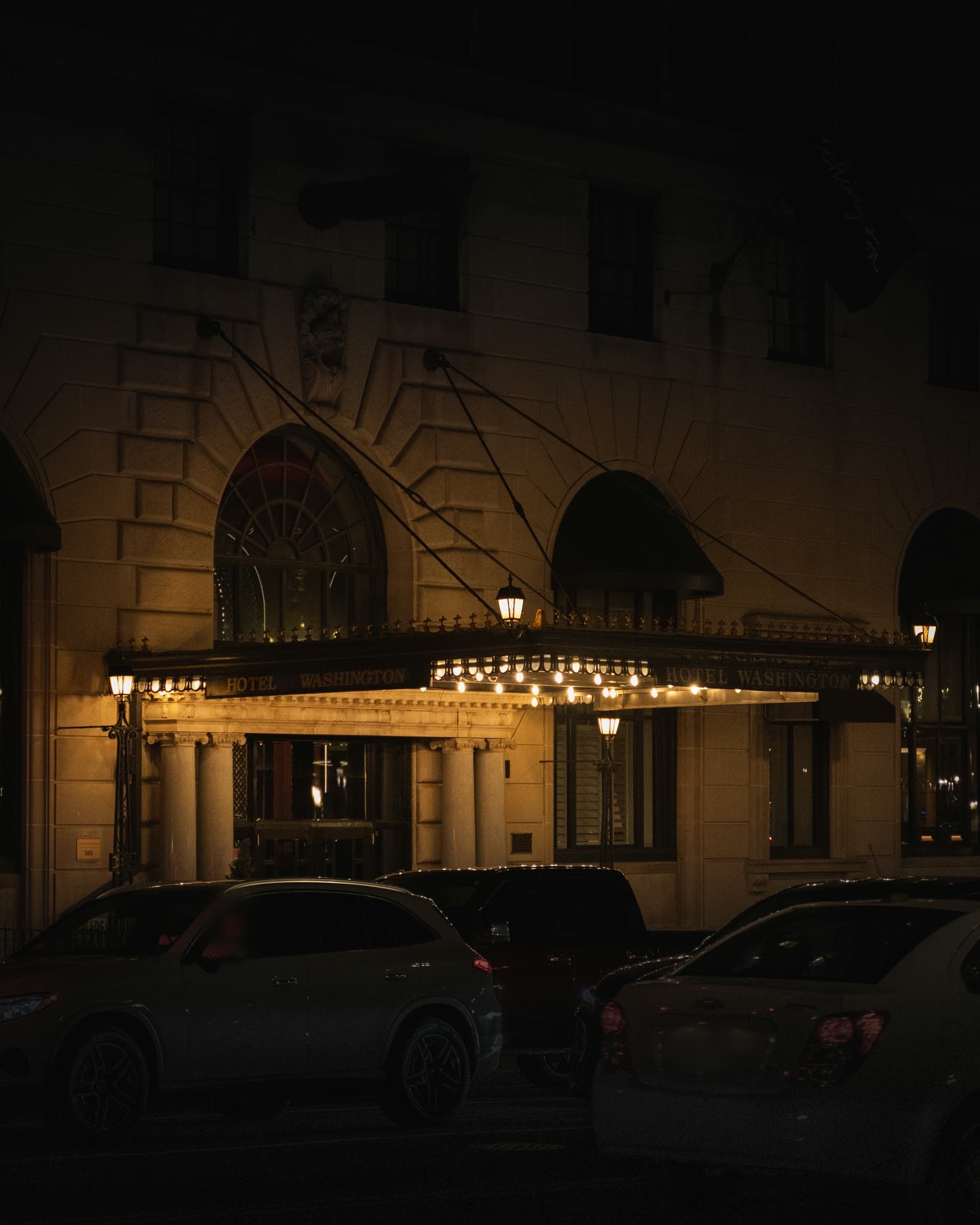 The lit canopy and lanterns of a historic hotel entrance at night, with cars at the curb