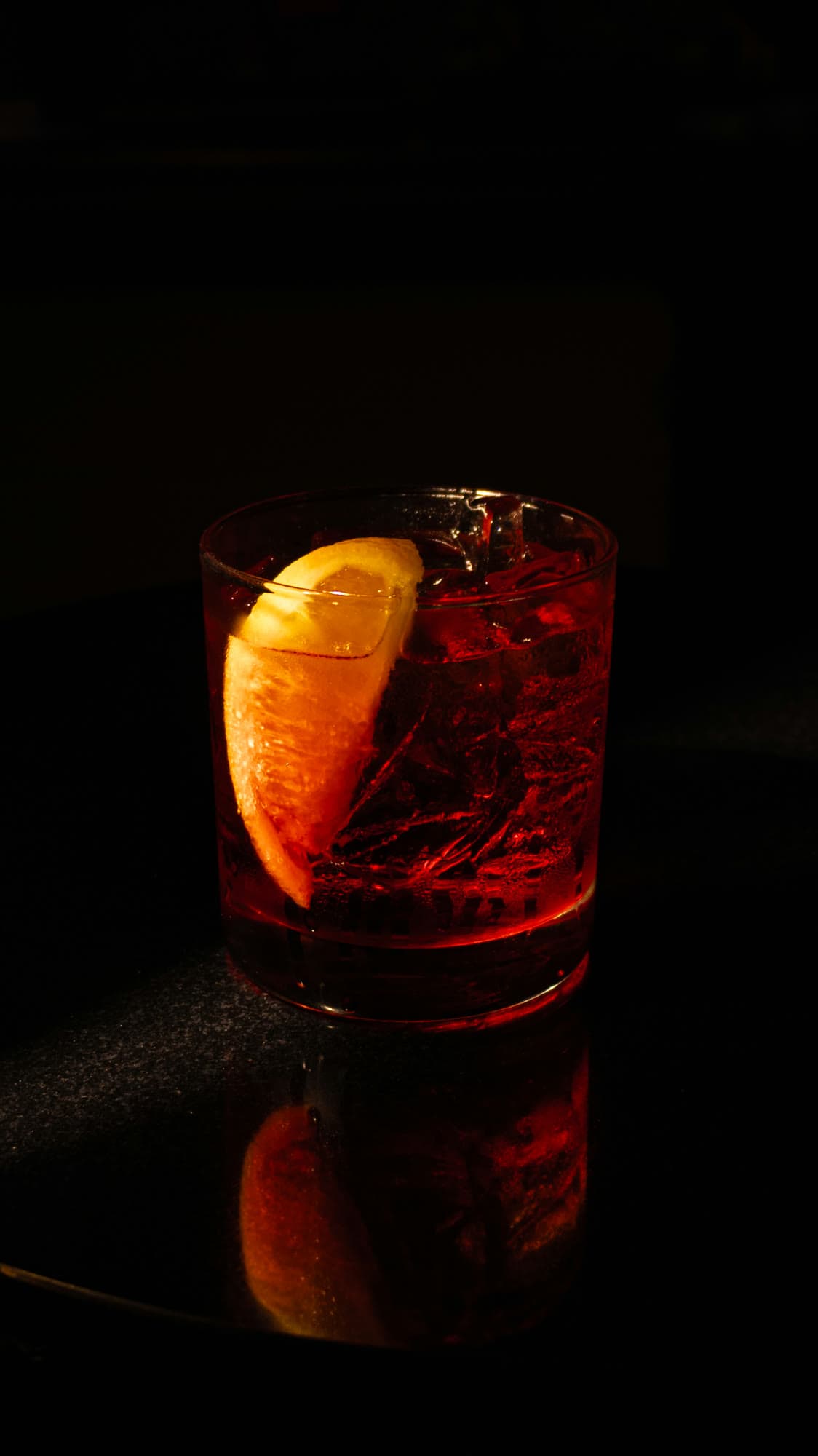 A red cocktail with an orange wedge, lit from one side against black