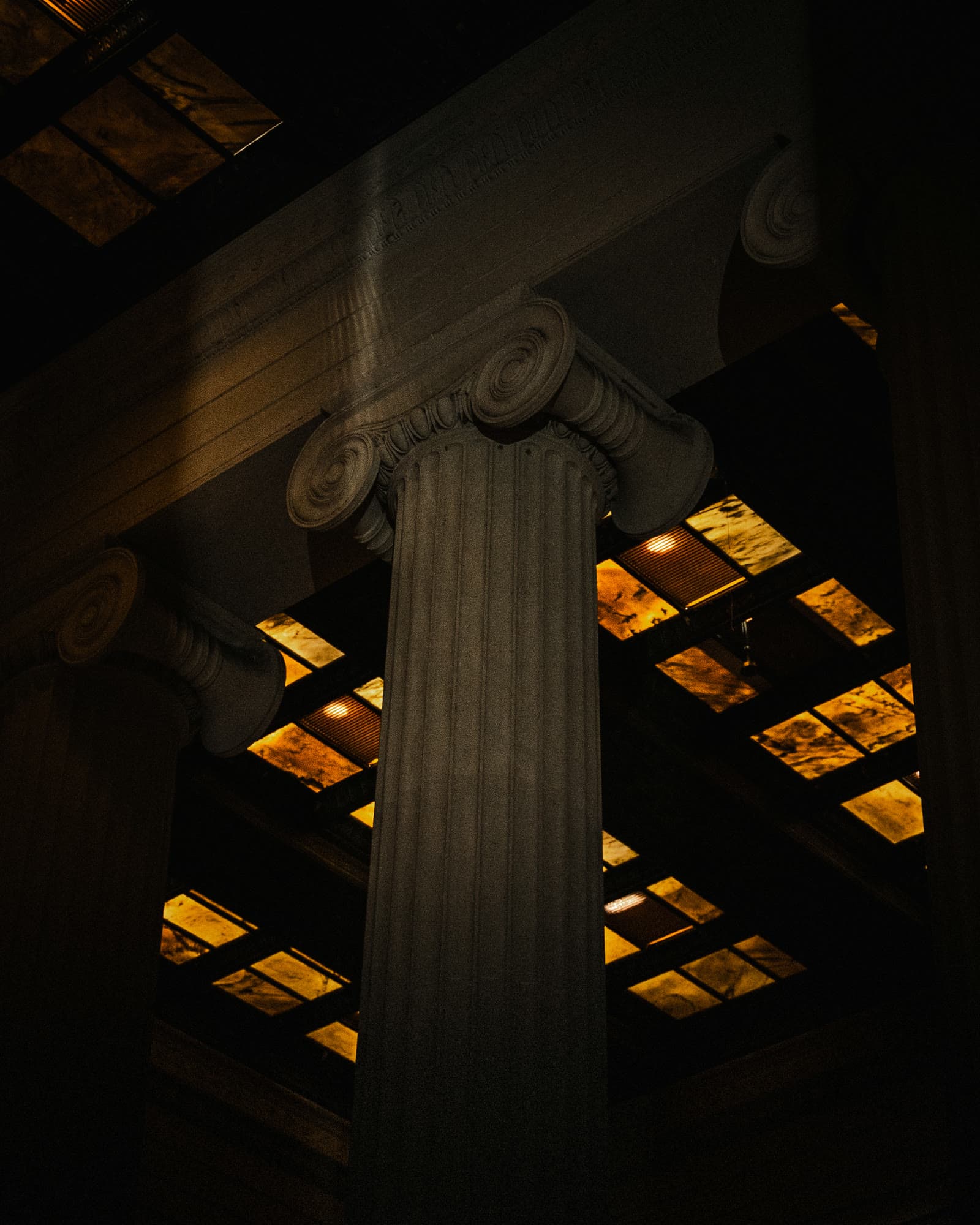An Ionic column rising toward a lit amber ceiling in a dark classical hall
