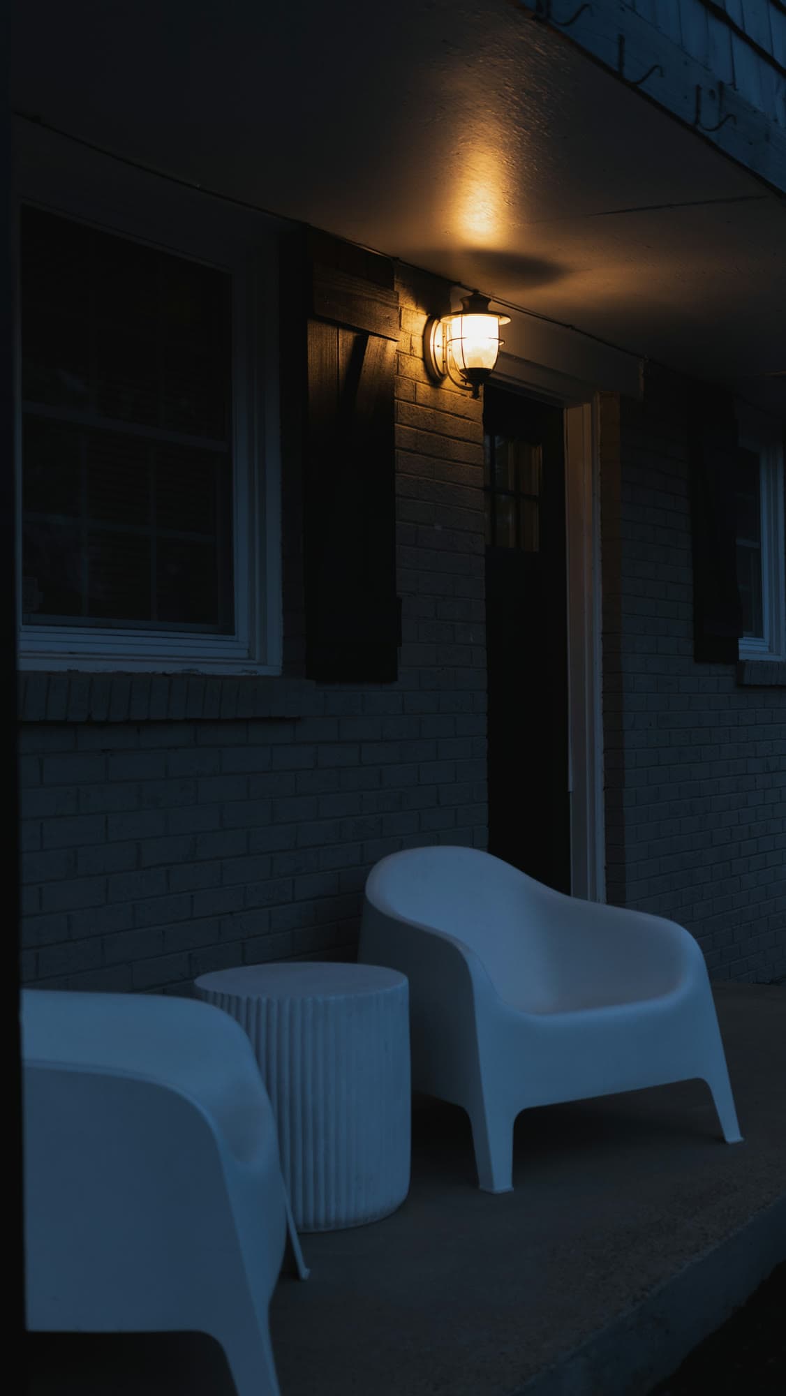 A porch lantern lighting a brick wall at blue hour above two white chairs