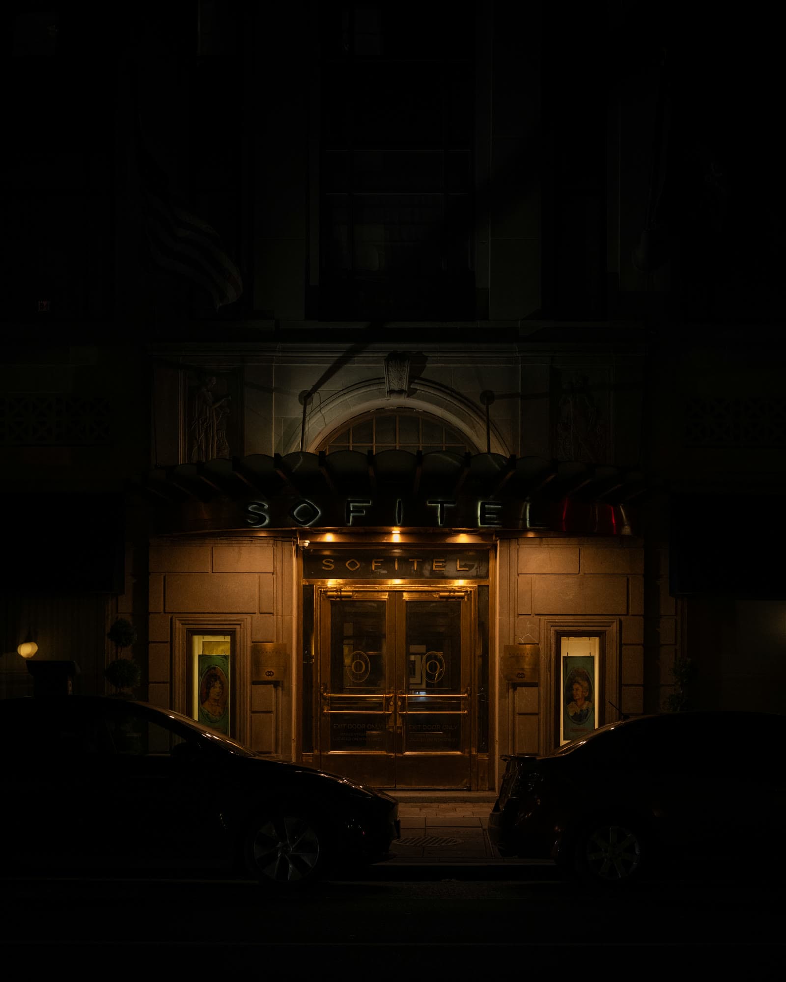 A hotel entrance glowing amber under its canopy at night, between two parked cars