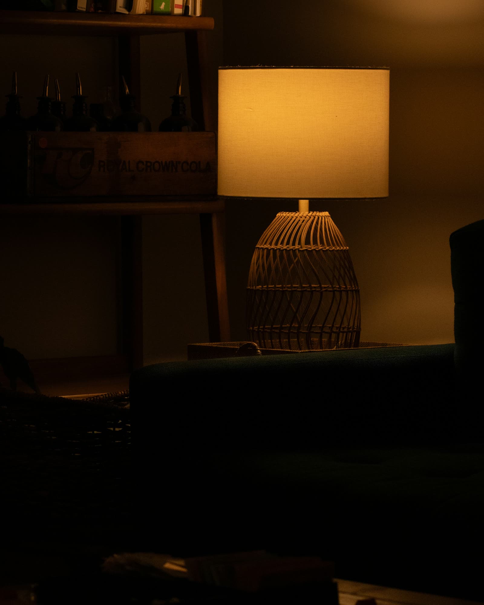 A table lamp with a woven base glowing in a dark room beside a shelf of bottles