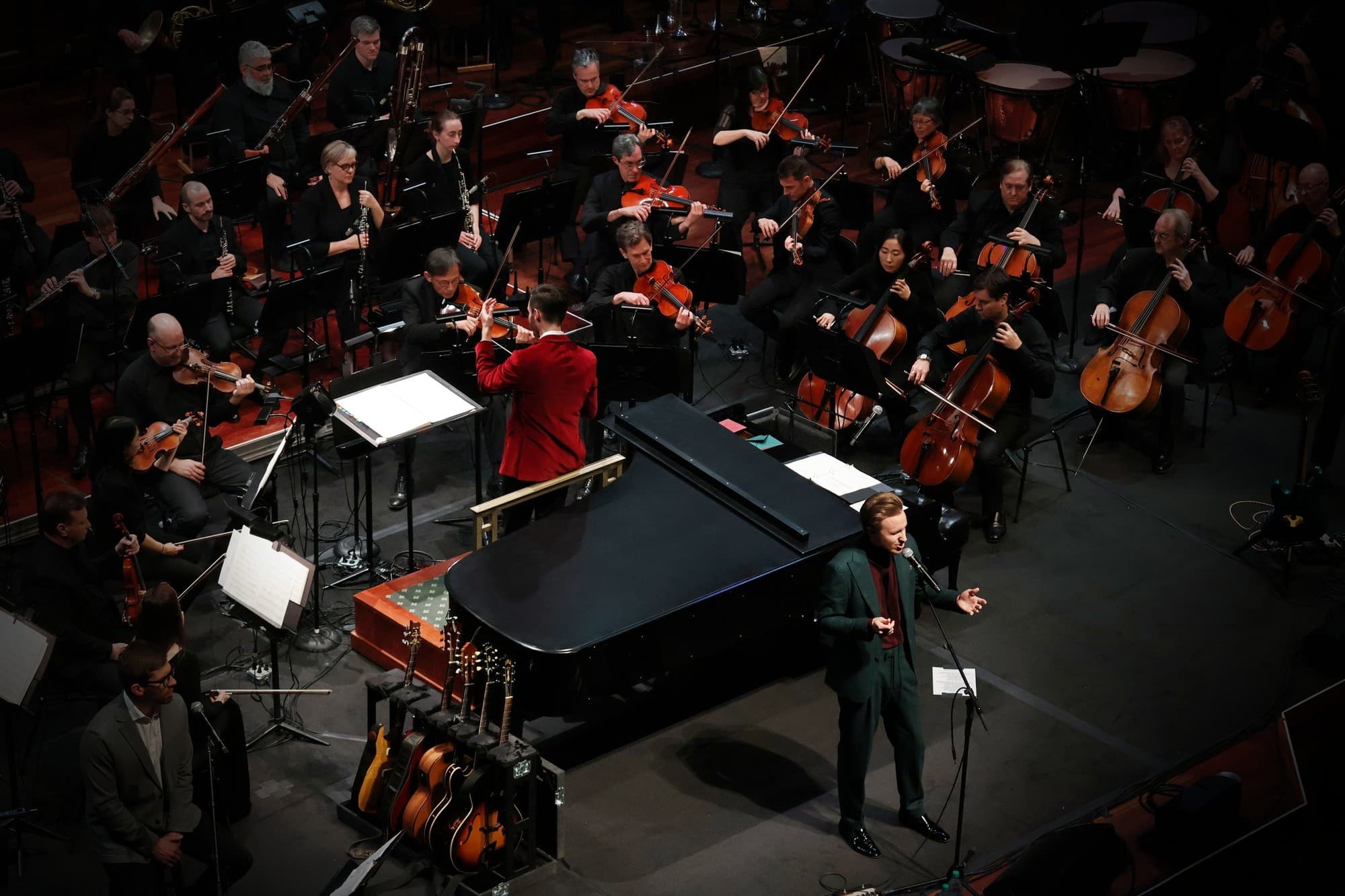 High angle view of the orchestra, the conductor in a red jacket and Cody Fry singing beside a grand piano, with guitars on stands in front