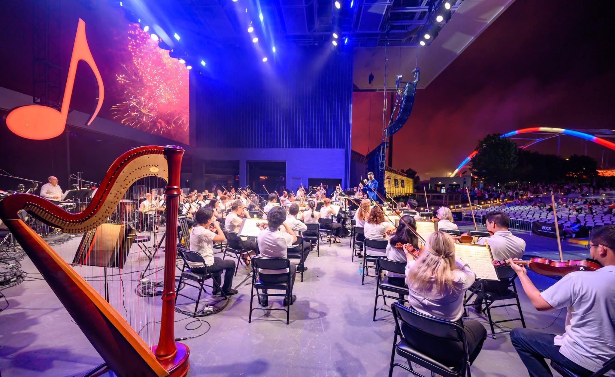 From behind a harp on the Ascend stage: the orchestra in white shirts, a conductor in a blue jacket at the right, fireworks on the LED wall beside a large orange musical note, and a lit arched bridge beyond the lawn