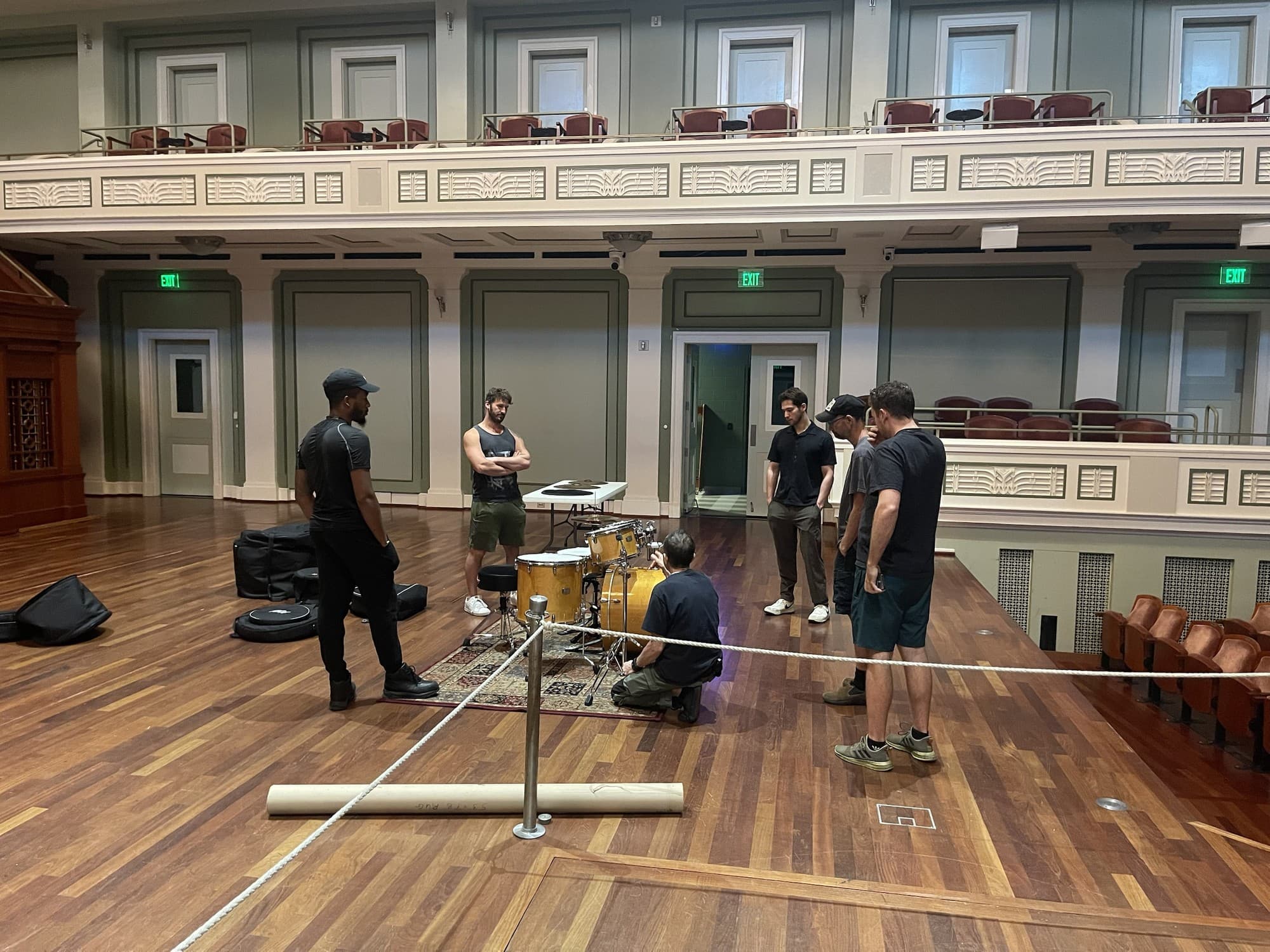 One stagehand kneeling at a drum kit while five others watch, with instrument cases and a rolled rug nearby