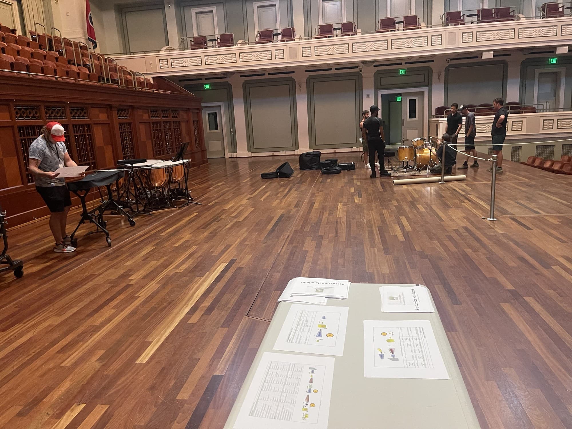Stagehands on the concert hall stage around a drum kit being set up, with printed handbook sheets on a table in the foreground